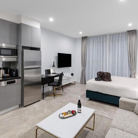 Meriton Suites Sussex Street, Sydney Eksteriør billede The photo shows a modern studio apartment. On one side, there is a kitchenette with sleek gray cabinets, a refrigerator, and built-in appliances. In the living area, there is a comfortable gray sofa, a white coffee table with a fruit platter and a bo