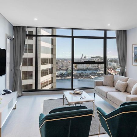 Meriton Suites Sussex Street, Sydney Eksteriør billede The photo shows a modern living room with large windows that offer a view of the cityscape and water. The interior features a light color palette with pale blue walls and soft lighting. There is a comfortable gray sofa and two green accent chairs, ar