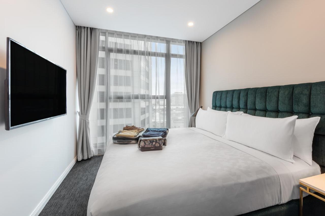 Meriton Suites Sussex Street, Sydney Eksteriør billede The photo shows a modern hotel bedroom. The room features a large bed with a green upholstered headboard and crisp white linens. There are two pillows on the bed and a few folded items of clothing neatly placed on it. A flat-screen TV is mounted on t