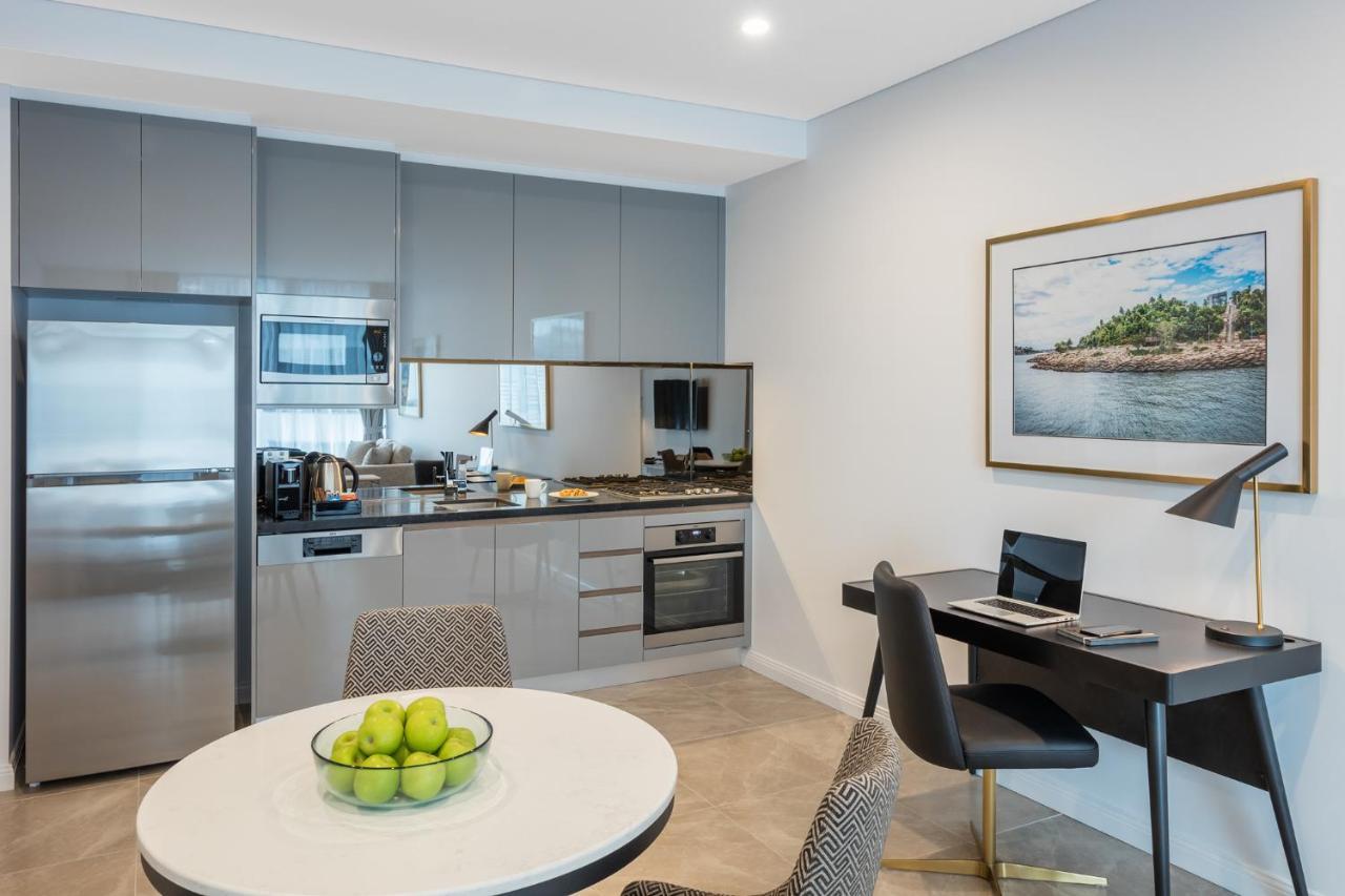 Meriton Suites Sussex Street, Sydney Eksteriør billede The photo shows a modern kitchen and dining area in an apartment. The kitchen features sleek gray cabinetry, a stainless steel fridge, and a microwave. There is a countertop with cooking appliances, including an oven and stovetop. A small round dinin