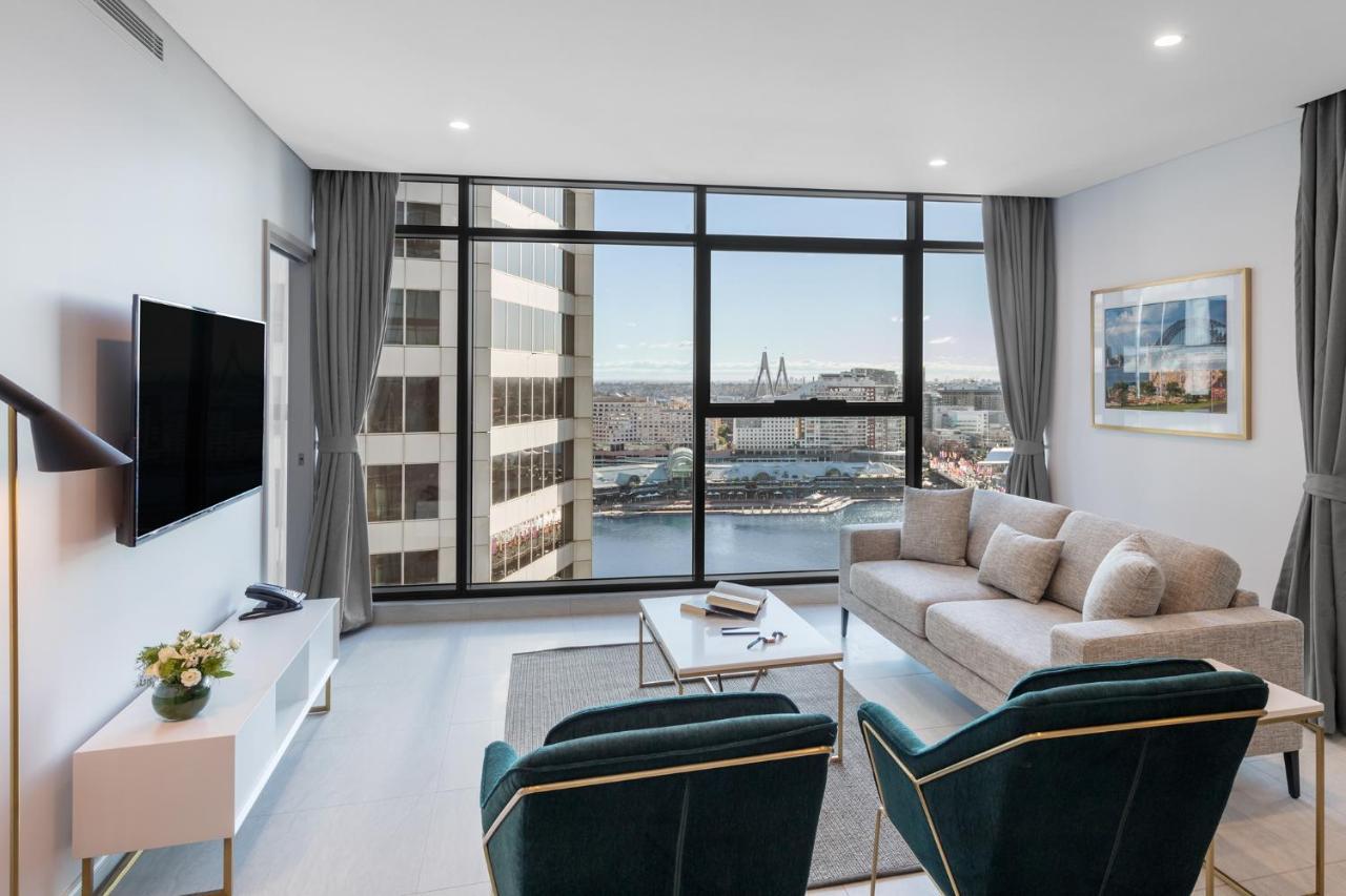Meriton Suites Sussex Street, Sydney Eksteriør billede The photo shows a modern living room with large windows that offer a view of the cityscape and water. The interior features a light color palette with pale blue walls and soft lighting. There is a comfortable gray sofa and two green accent chairs, ar