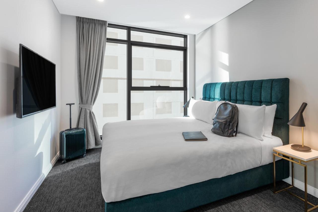 Meriton Suites Sussex Street, Sydney Eksteriør billede The photo shows a modern hotel room featuring a comfortable bed with white bedding and a teal-colored headboard. There is a neatly placed black backpack on the bed, along with a small notebook or tablet. A television is mounted on the wall opposite t