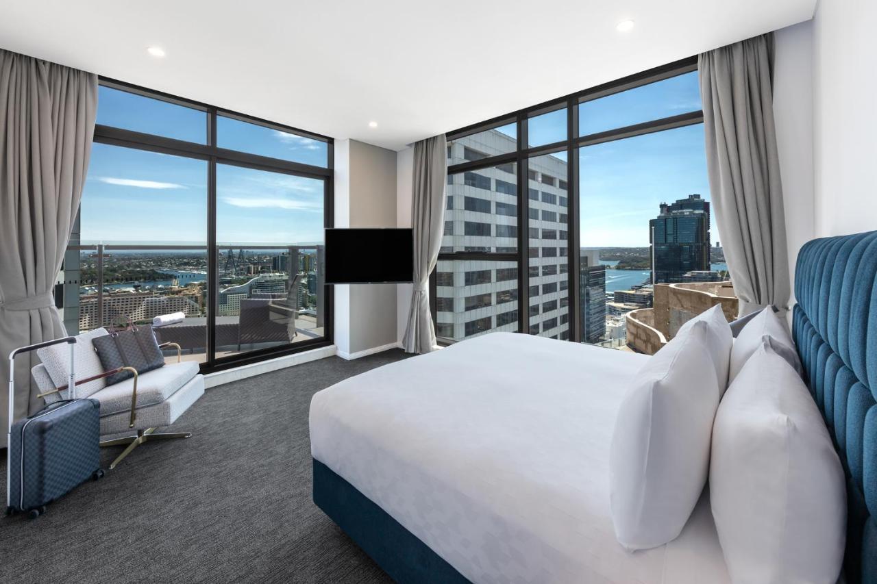 Meriton Suites Sussex Street, Sydney Eksteriør billede The photo shows a modern hotel room featuring large windows that provide a panoramic view of the cityscape and waterfront. The room is furnished with a king-size bed dressed in white linens, a comfortable armchair, and a flat-screen TV mounted on the