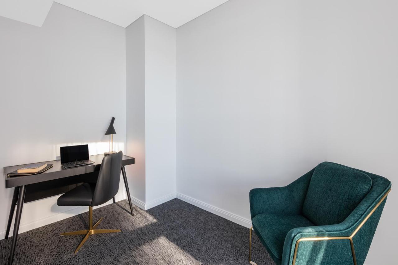 Meriton Suites Sussex Street, Sydney Eksteriør billede The photo depicts a minimalist interior space, likely a small office or study area. It features a simple, modern desk with a black lamp and a laptop or tablet on it. A sleek black chair accompanies the desk, while on the opposite side of the room, th