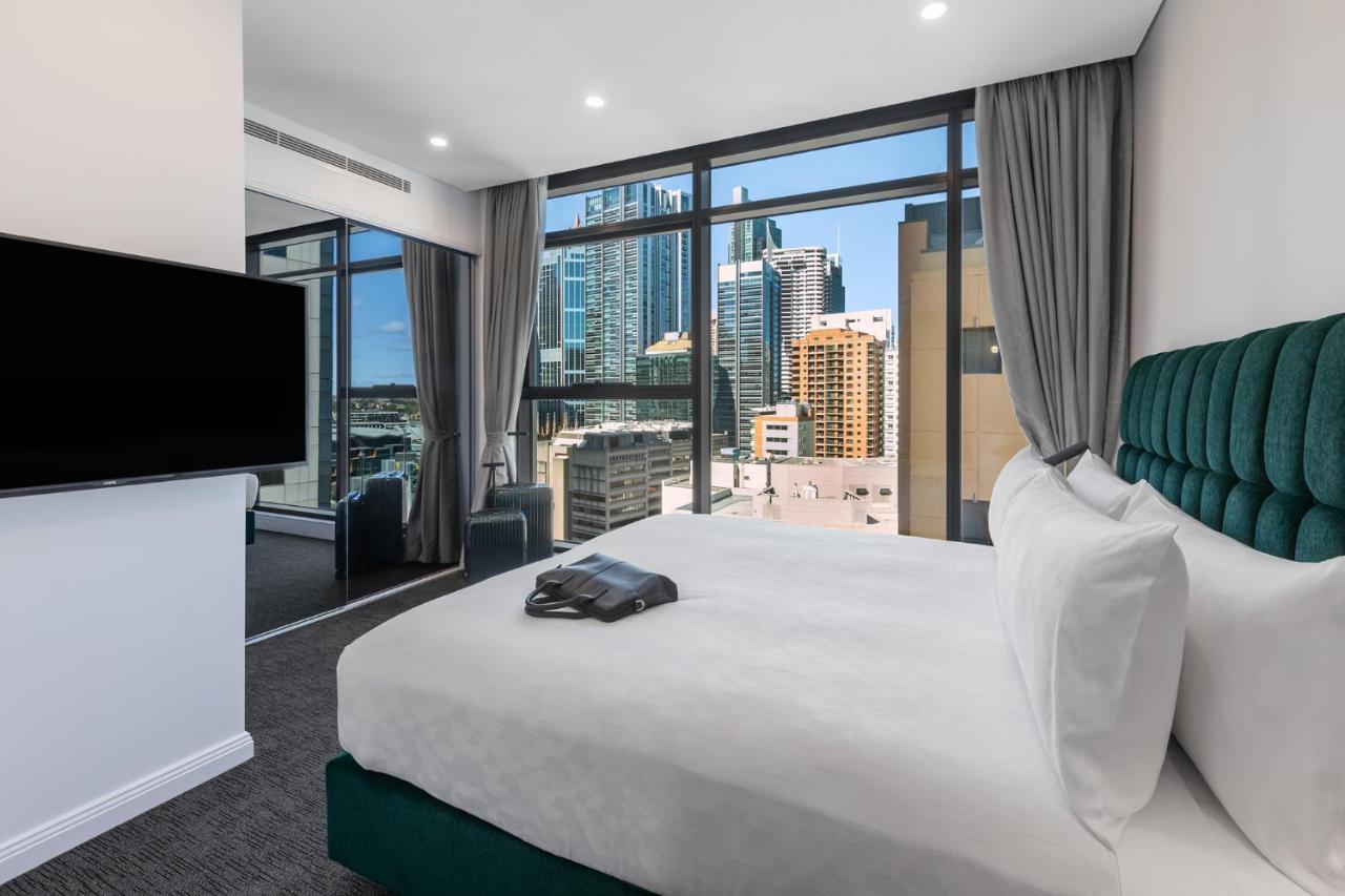 Meriton Suites Sussex Street, Sydney Eksteriør billede The photo shows a modern hotel room featuring a large bed with white linens and a green upholstered headboard. There is a sleek television mounted on the wall opposite the bed, and the room has large windows that provide a view of a cityscape, includ