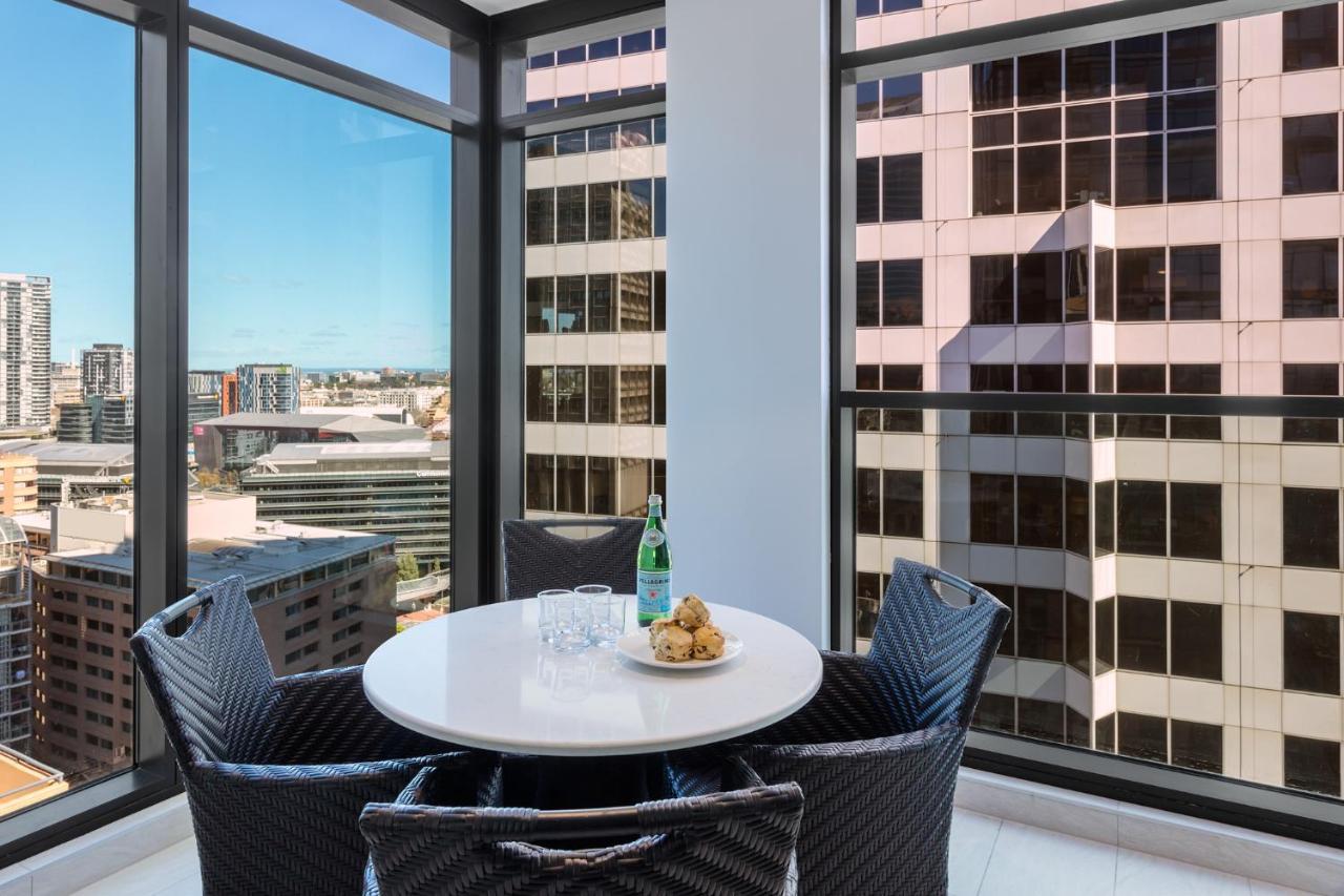 Meriton Suites Sussex Street, Sydney Eksteriør billede The photo shows a modern dining space with a round white table set for four. There are four black woven chairs around the table. On the table, there's a bottle of sparkling water, two glasses, and a small plate with several cookies. The large windows