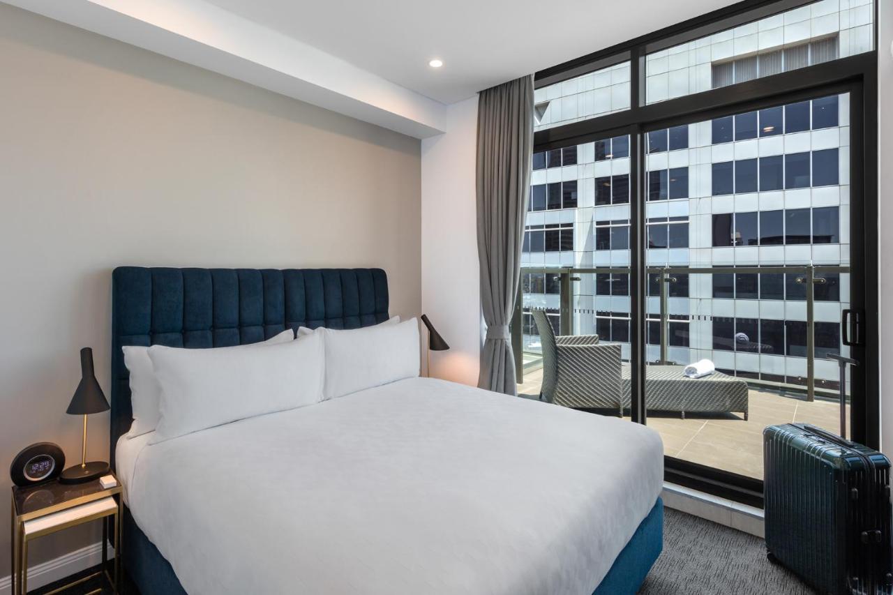 Meriton Suites Sussex Street, Sydney Eksteriør billede The photo shows a modern hotel room with a large bed featuring a blue upholstered headboard and white linens. Natural light enters through a large window, revealing a view of a building exterior, likely part of the hotel or surrounding area. The room