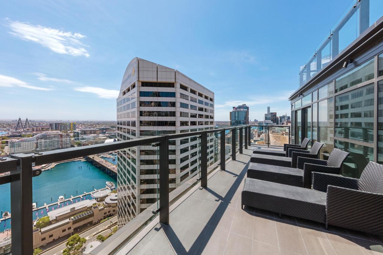 Meriton Suites Sussex Street, Sydney Eksteriør billede The photo shows a balcony or terrace with a panoramic view of a city skyline. The balcony features modern outdoor furniture, including sleek lounge chairs. In the background, you can see a waterfront area with buildings and a harbor, along with a cle