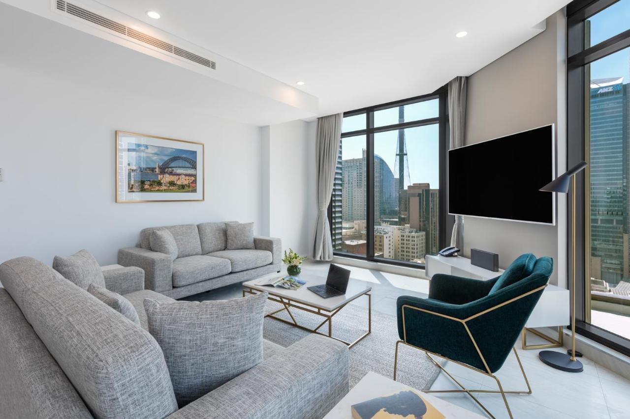 Meriton Suites Sussex Street, Sydney Eksteriør billede The photo shows a modern living room with large windows offering a view of a city skyline. The room features two gray sofas and a stylish green accent chair. A sleek coffee table is placed in front of the sofas, alongside a laptop. There’s a flat-scr