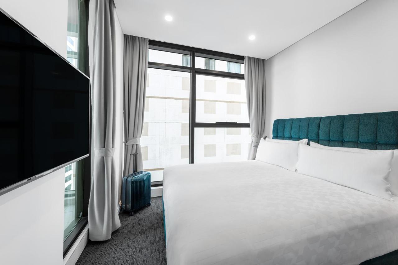 Meriton Suites Sussex Street, Sydney Eksteriør billede The photo shows a modern hotel room featuring a large bed with a light-colored duvet and a blue upholstered headboard. There are two pillows on the bed. A large window allows natural light to enter the room, and gray curtains frame the window. Next t