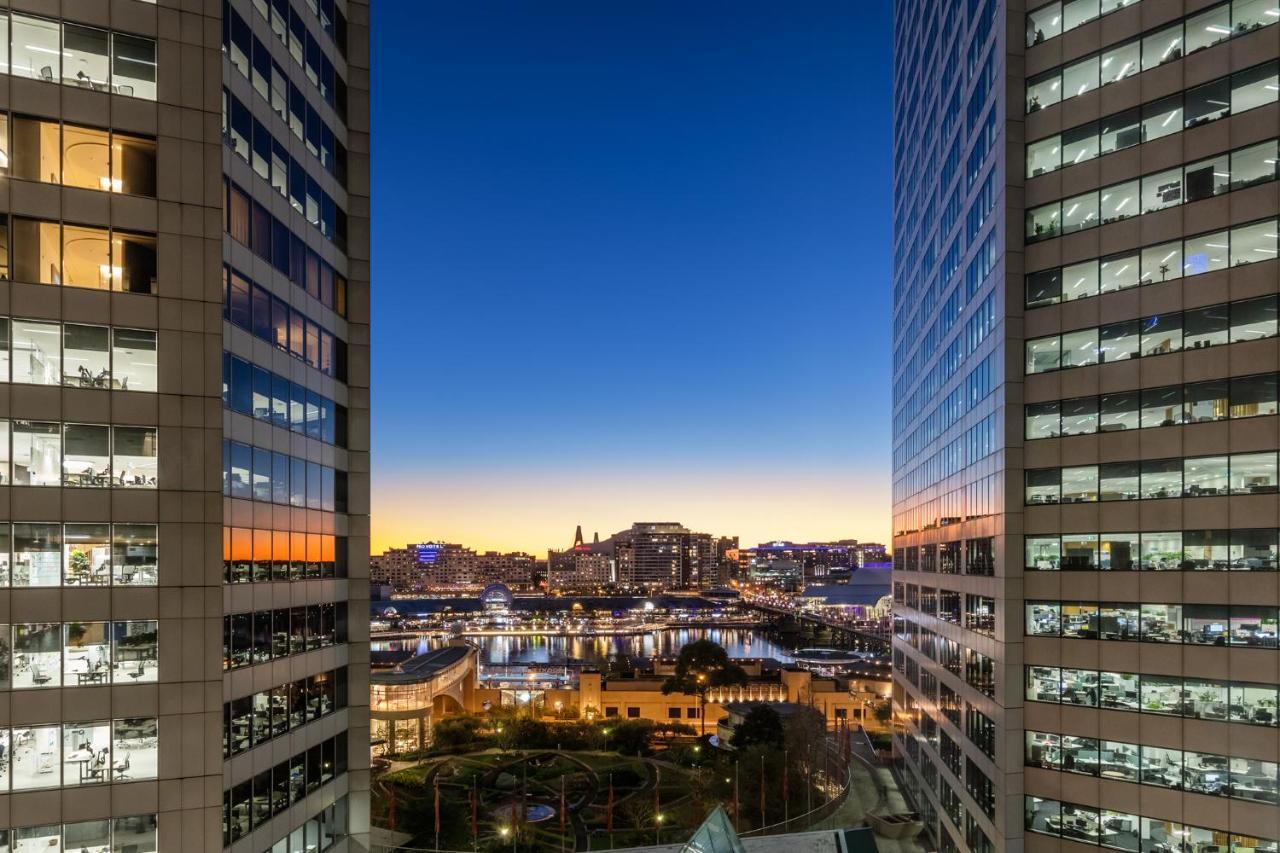 Meriton Suites Sussex Street, Sydney Eksteriør billede The photo shows a view between two tall buildings, likely office towers, during twilight. On one side, there are glass windows reflecting the evening sky that transitions from blue to orange near the horizon. In the background, a cityscape is visible