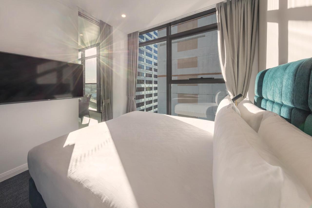 Meriton Suites Sussex Street, Sydney Eksteriør billede The photo shows a modern hotel room with a neatly made bed featuring white bedding and a plush, tufted headboard in a shade of blue. Natural light is streaming through large windows, which are covered partially by light gray curtains. A flat-screen t