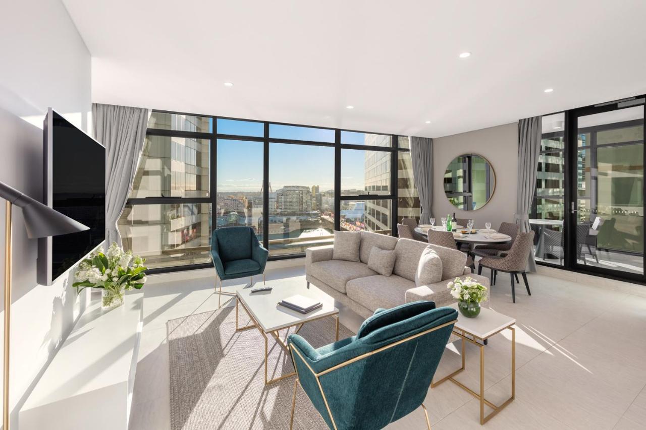Meriton Suites Sussex Street, Sydney Eksteriør billede The photo shows a modern and stylish living space. It features a spacious room with large windows that provide a bright view of the cityscape. The furniture includes a light gray sofa, two teal chairs, and a coffee table. There is a flat-screen TV mo