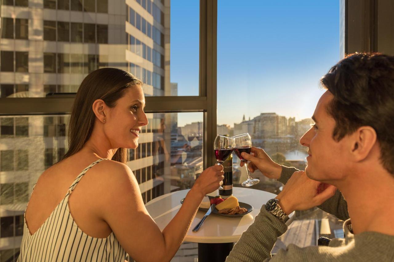 Meriton Suites Sussex Street, Sydney Eksteriør billede The photo depicts a couple enjoying a drink together in a modern setting. They are sitting at a small table near a large window, which provides a view of a cityscape and a sunset. Both individuals are smiling and engaged in a cheerful toast with glas