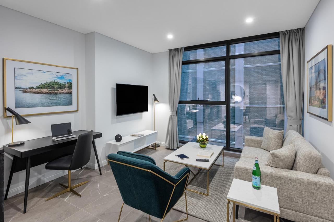Meriton Suites Sussex Street, Sydney Eksteriør billede The photo shows a modern living area with a light color scheme. There is a comfortable seating arrangement that includes a beige sofa and an accent chair in teal. A coffee table is positioned in front of the sofa, with a small vase of flowers on it. 