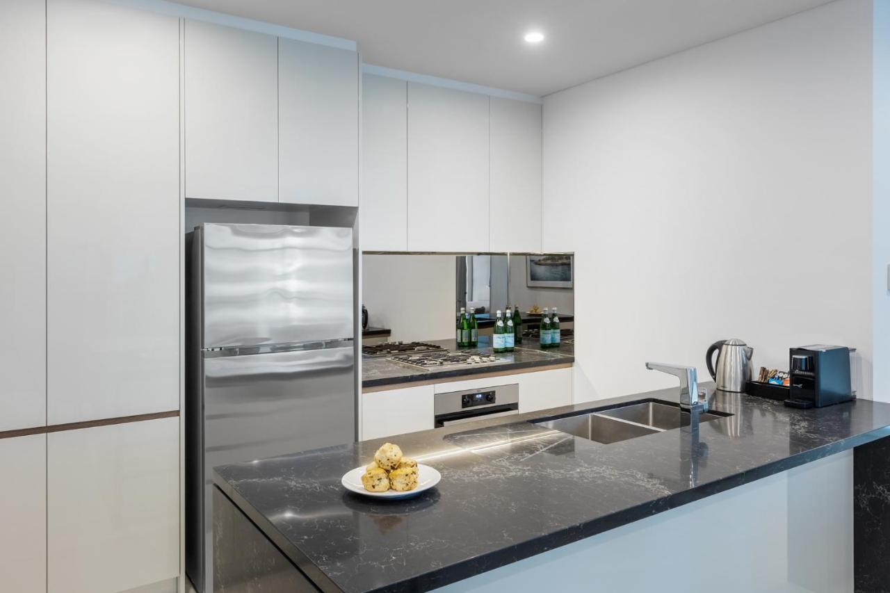 Meriton Suites Sussex Street, Sydney Eksteriør billede The photo shows a modern kitchen with a sleek, minimalist design. Key features include:

- A shiny stainless steel refrigerator on the left.
- A mirrored backsplash above the stove, which has a gas burner.
- A dark countertop made of what appears to 