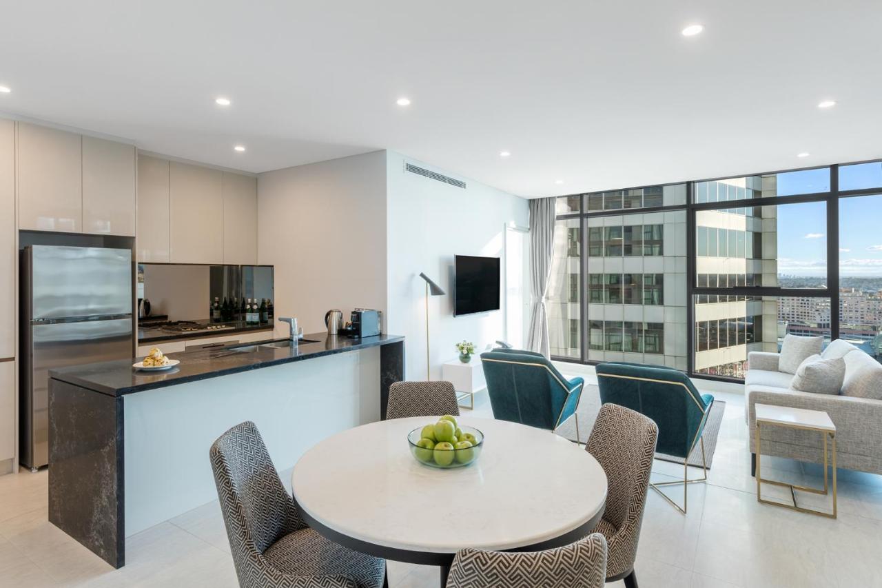 Meriton Suites Sussex Street, Sydney Eksteriør billede The photo shows a modern and stylish interior space, likely a living room and kitchen area. The kitchen features sleek cabinetry and a countertop, with appliances visible, including a coffee maker. There is a round dining table with gray chairs, set 