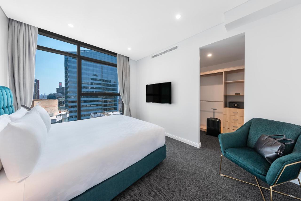 Meriton Suites Sussex Street, Sydney Eksteriør billede The photo shows a modern hotel room. It features a large window that allows natural light to fill the space, offering a view of the city skyline. The room includes a neatly made bed with a white duvet and a teal-colored base. There is a small green a