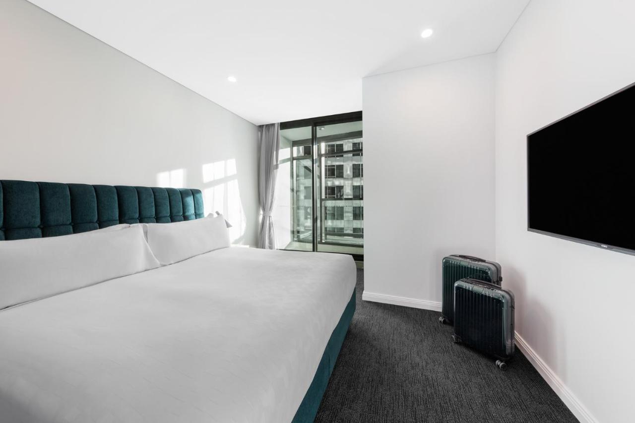 Meriton Suites Sussex Street, Sydney Eksteriør billede The photo shows a modern hotel room featuring a large bed with a headboard in a teal color. The bedding is white and appears neatly arranged. There is a large window allowing natural light to enter, offering a view of an outdoor area. A flat-screen T