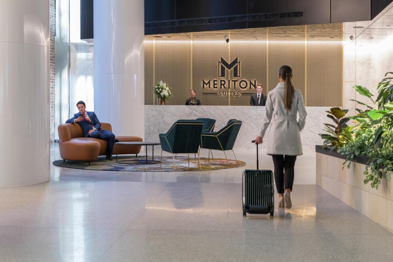 Meriton Suites Sussex Street, Sydney Eksteriør billede The photo depicts a modern hotel lobby. In the foreground, a woman is walking towards the reception desk, pulling a suitcase behind her. She is dressed in a light-colored coat. To her left, there’s a comfortable seating area with a person sitting on 