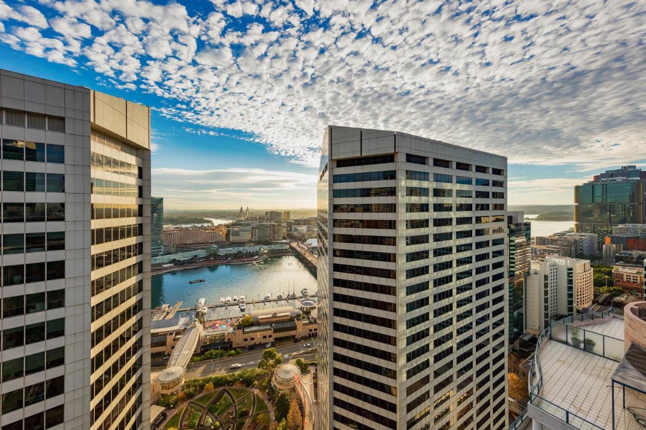 Meriton Suites Sussex Street, Sydney Eksteriør billede The photo depicts a panoramic view of a modern cityscape featuring tall commercial buildings with glass facades. The shot is taken from a height, showing the intricate architecture of the nearby structures. Below, there is a body of water, which appe