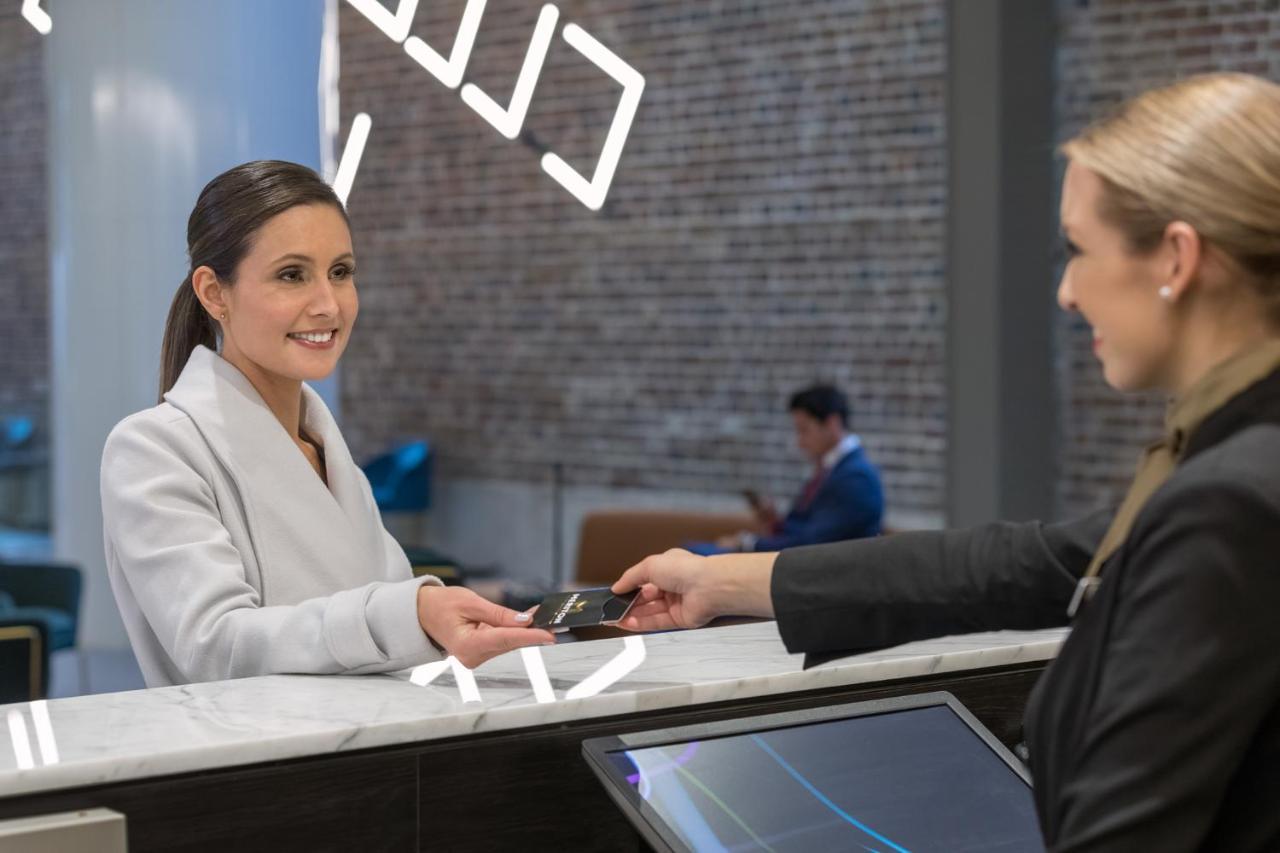 Meriton Suites Sussex Street, Sydney Eksteriør billede The image depicts a scene at a hotel reception desk. A woman with long hair, dressed in a white outfit, is smiling as she hands her card to a receptionist, who is in a black uniform. The reception area features a modern design, and there is a person 
