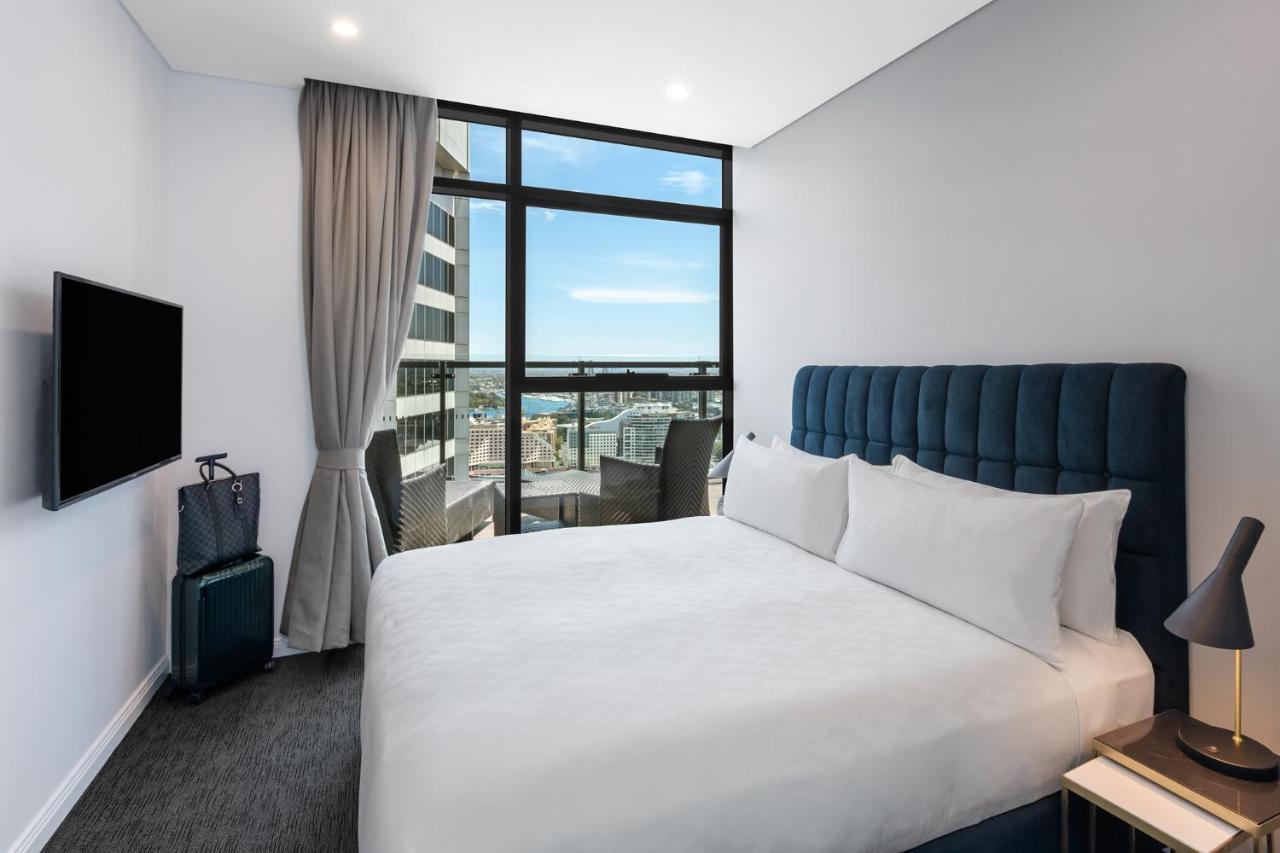Meriton Suites Sussex Street, Sydney Eksteriør billede The photo shows a modern hotel room featuring a double bed with white linens and a tufted blue headboard. There is a small bedside table next to the bed. A large window allows natural light to fill the room, offering a view of the city outside, inclu