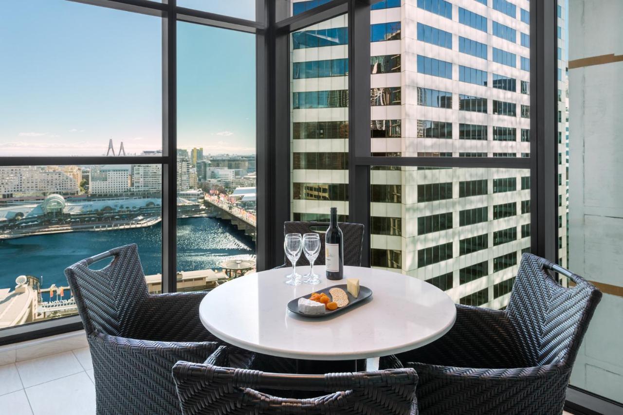 Meriton Suites Sussex Street, Sydney Eksteriør billede The photo shows a modern dining area with a round table and four woven chairs. On the table, there are two wine glasses, a bottle of wine, and a plate with food items. The large windows provide a view of a cityscape featuring tall buildings and a bod