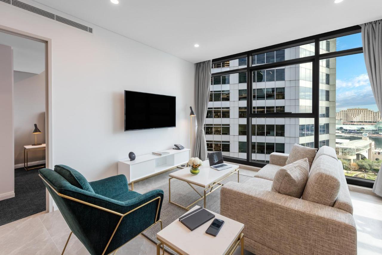 Meriton Suites Sussex Street, Sydney Eksteriør billede The photo shows a modern living room with large windows that let in natural light, providing a view of a cityscape outside. The interior features a comfortable light-colored sofa and a stylish dark green armchair. A sleek TV is mounted on the wall op