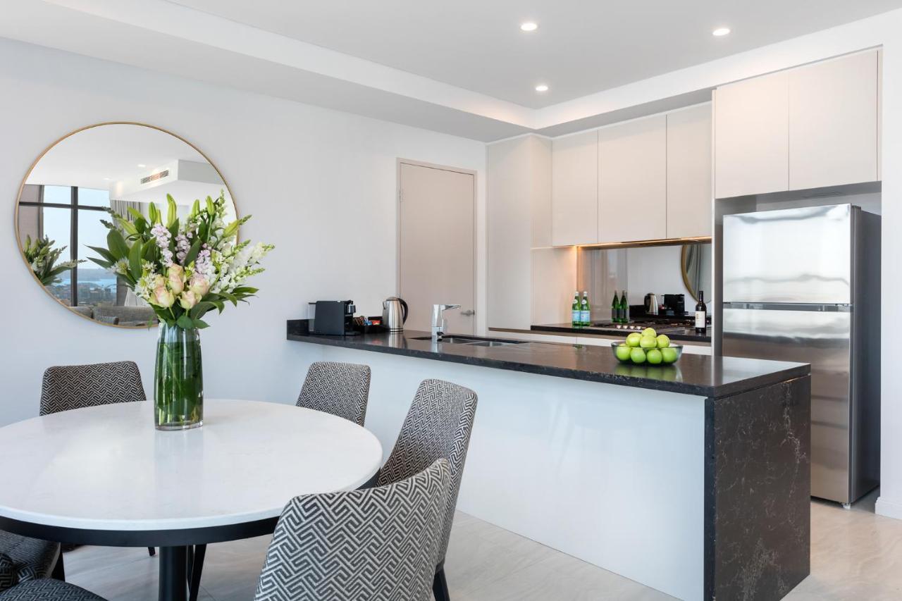 Meriton Suites Sussex Street, Sydney Eksteriør billede The photo shows a modern kitchen and dining area. In the foreground, there's a circular dining table surrounded by four stylish chairs. A large vase filled with fresh flowers is placed on the table, adding a touch of color and elegance. 

In the kitc