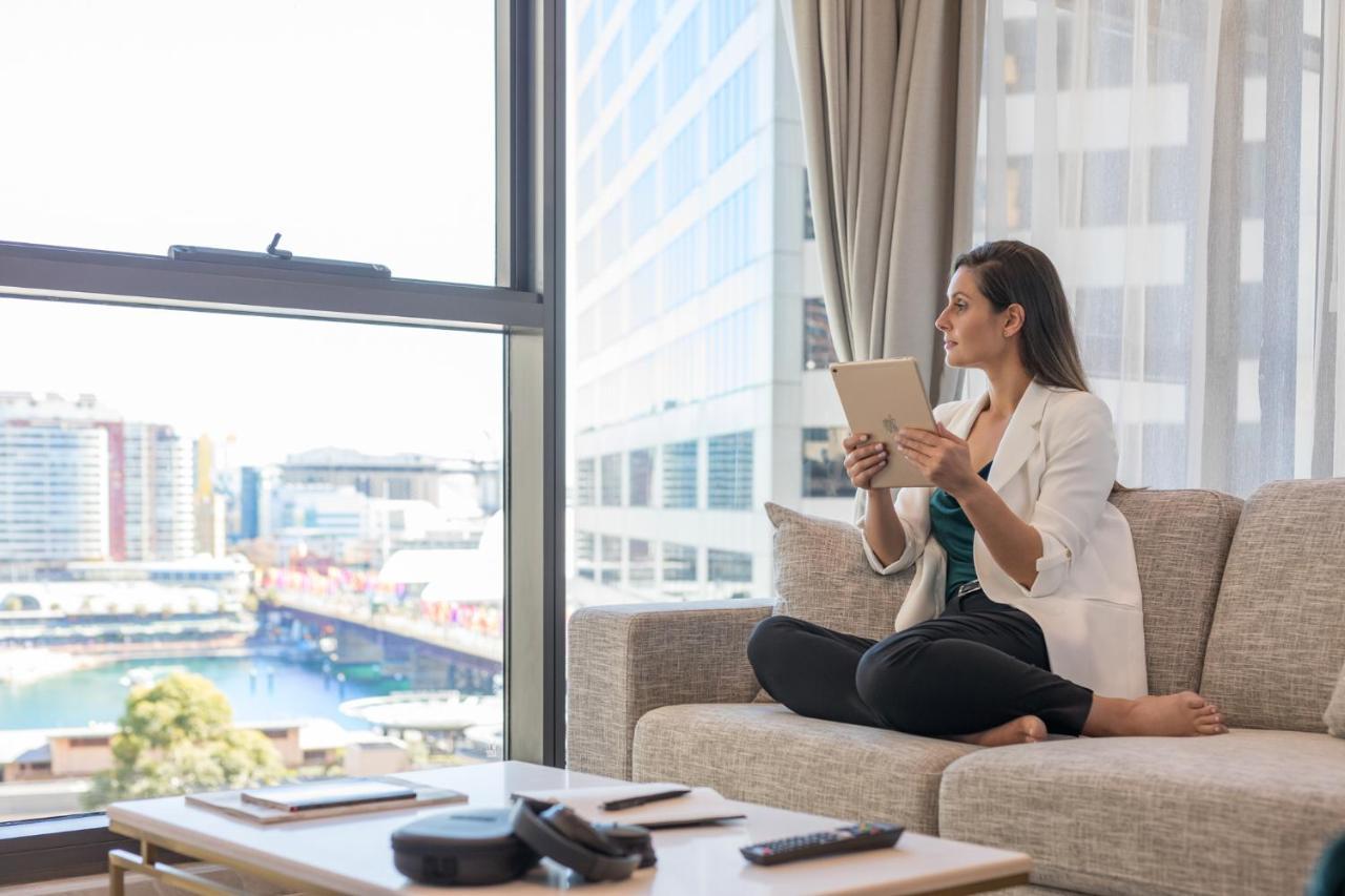 Meriton Suites Sussex Street, Sydney Eksteriør billede The image portrays a woman sitting cross-legged on a light-colored sofa, engaged with a tablet in her hands. She is dressed in a white blazer and dark pants, appearing relaxed and focused. The setting features large windows that provide a view of a w