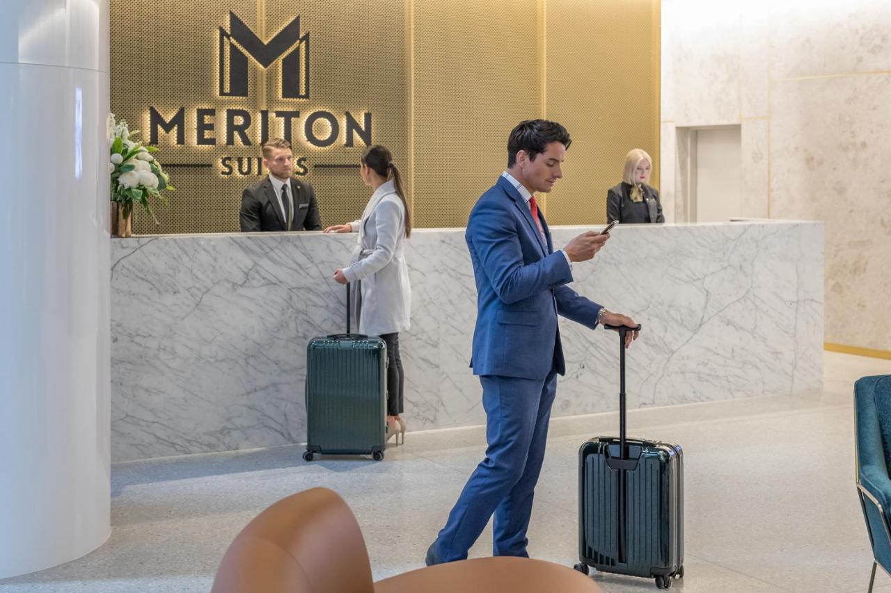 Meriton Suites Sussex Street, Sydney Eksteriør billede The photo shows the interior of a hotel reception area, specifically at Meriton Suites. In the foreground, a man in a blue suit is standing with a suitcase, looking at his phone. Behind him, there are two staff members at the reception desk, one of w