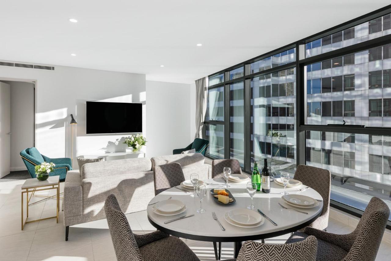 Meriton Suites Sussex Street, Sydney Eksteriør billede The photo shows a modern and stylish living room and dining area. In the foreground, there is a round dining table set for a meal with plates, cutlery, and drinks. The seating arrangement includes comfortable chairs around the table. 

In the backgro