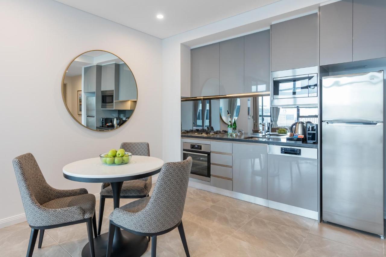 Meriton Suites Sussex Street, Sydney Eksteriør billede The photo features a modern kitchen and dining area. The kitchen has sleek grey cabinets and countertops, with built-in appliances including an oven and possibly a cooktop. There's a large stainless steel refrigerator on the right. A round dining tab