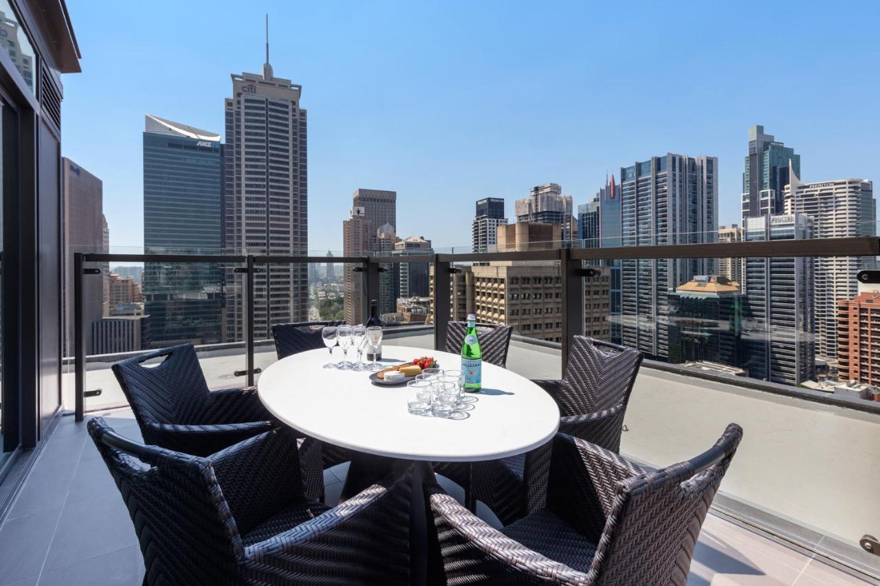 Meriton Suites Sussex Street, Sydney Eksteriør billede The photo shows a stylish outdoor dining area on a balcony with a round white table surrounded by black wicker chairs. On the table, there are several glasses, a bottle of sparkling water, and a plate of snacks. In the background, there is a panorami
