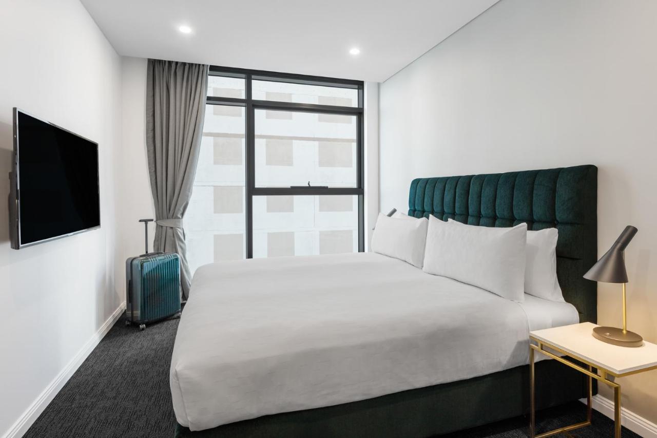 Meriton Suites Sussex Street, Sydney Eksteriør billede The photo shows a modern hotel room. It features a large bed with a dark green tufted headboard and multiple white pillows. There's a nightstand with a lamp beside the bed. A sleek television is mounted on the wall opposite the bed, and a suitcase is