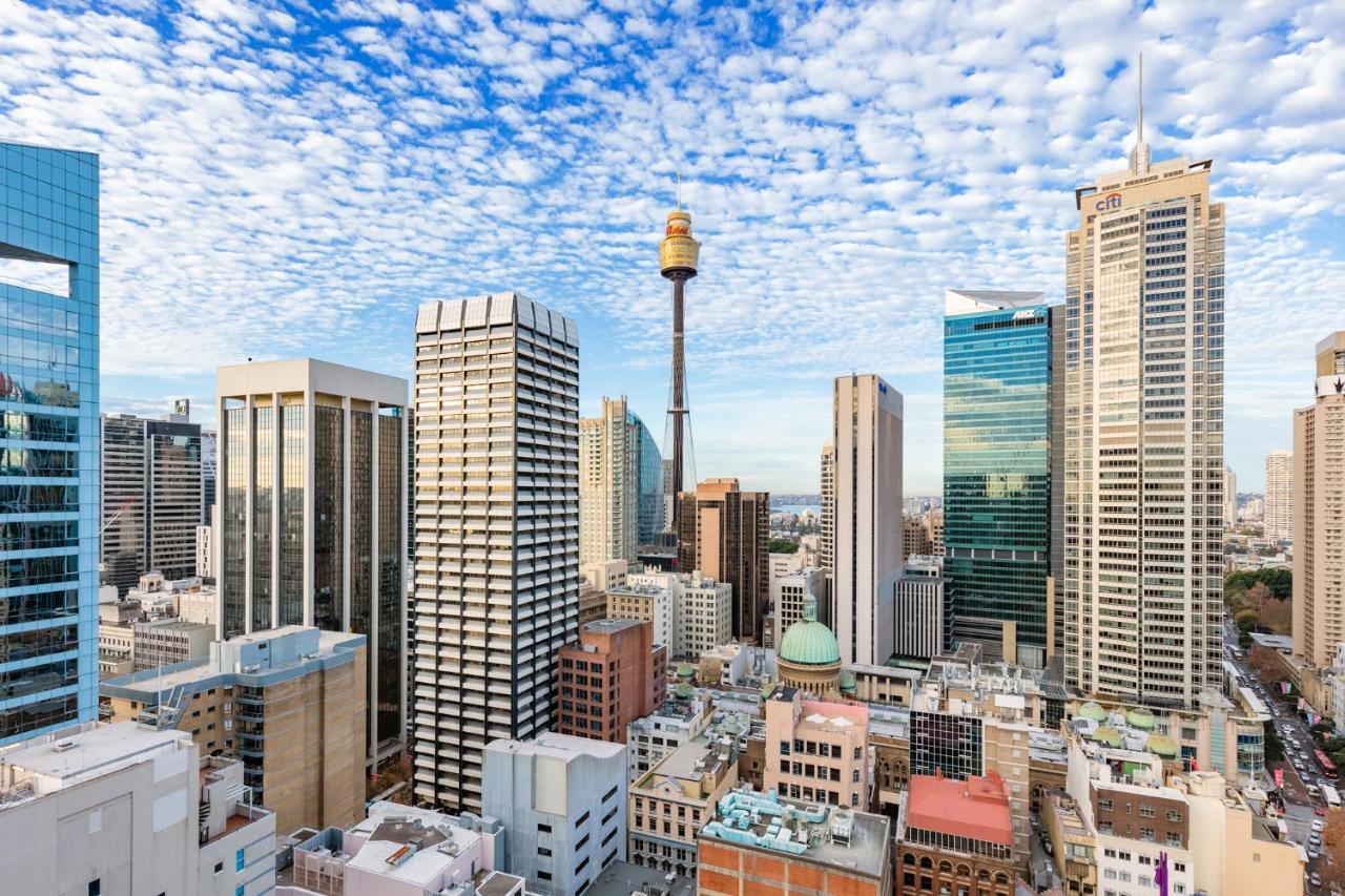 Meriton Suites Sussex Street, Sydney Eksteriør billede The photo shows a panoramic view of a city skyline dominated by tall skyscrapers. In the center, there is a notable tower with a distinctive golden structure at its top, which is likely a landmark of the city. The buildings have a mix of modern glass