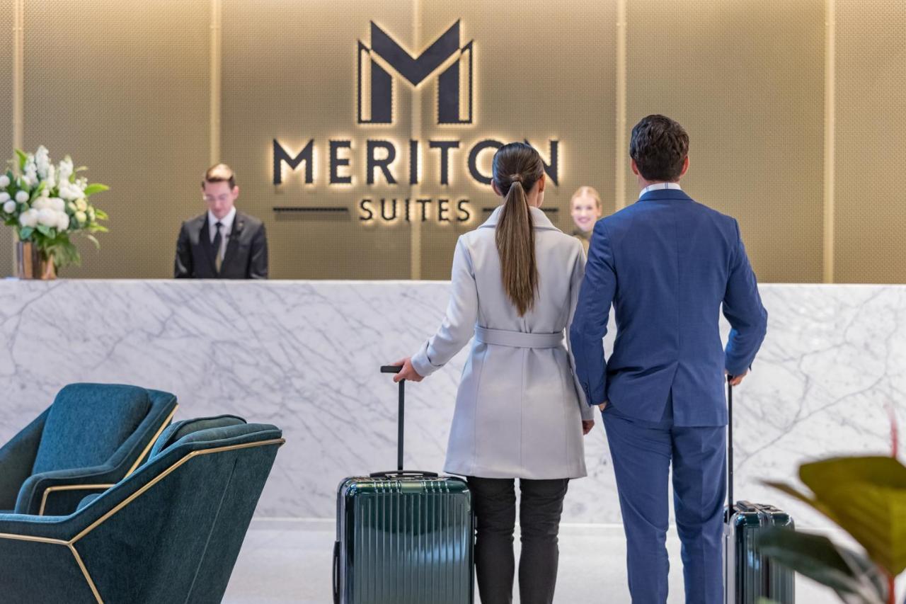 Meriton Suites Sussex Street, Sydney Eksteriør billede The photo shows a couple standing at a reception area in a hotel named "Meriton Suites." They are both facing the reception desk, where staff members are visible. The man is dressed in a suit, and the woman is wearing a light-colored coat. They each 