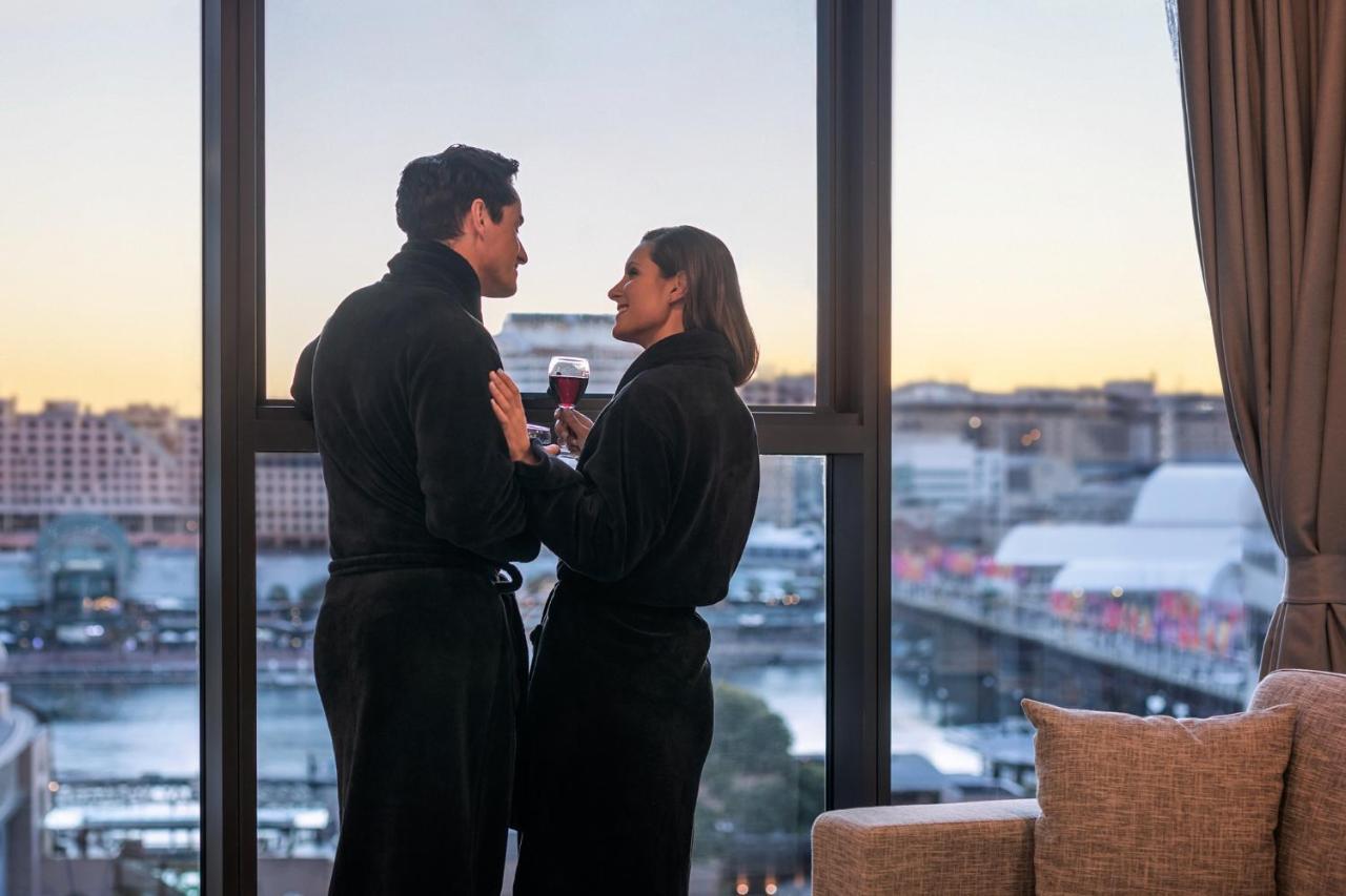 Meriton Suites Sussex Street, Sydney Eksteriør billede The photo depicts a couple standing closely together by a large window. They are wearing dark bathrobes and holding drinks, gazing into each other’s eyes. The background features a scenic view, likely of a cityscape at sunset, with soft light illumin