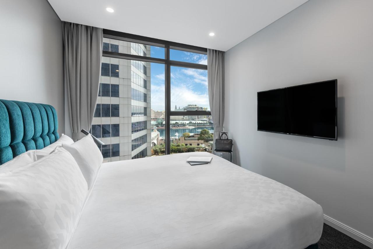 Meriton Suites Sussex Street, Sydney Eksteriør billede The photo shows a modern hotel room with a large bed featuring a white comforter and pillows. There is a blue headboard that adds a pop of color to the decor. A large window provides a view of the outside, which includes buildings and a glimpse of wa