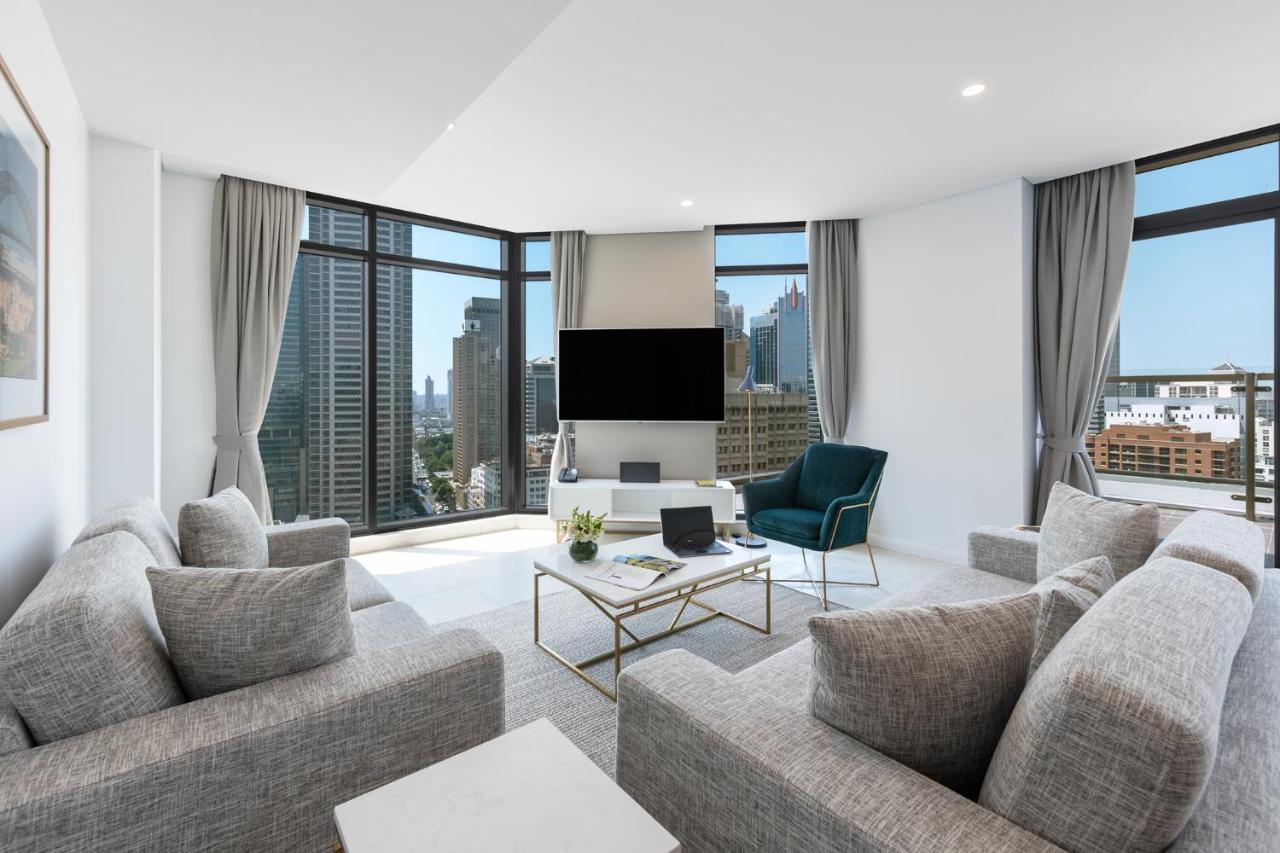 Meriton Suites Sussex Street, Sydney Eksteriør billede The photo showcases a modern living room with large windows offering a view of a city skyline. The room features a light color palette with gray sofas arranged around a coffee table. A flat-screen TV is mounted on the wall, and there is a green armch