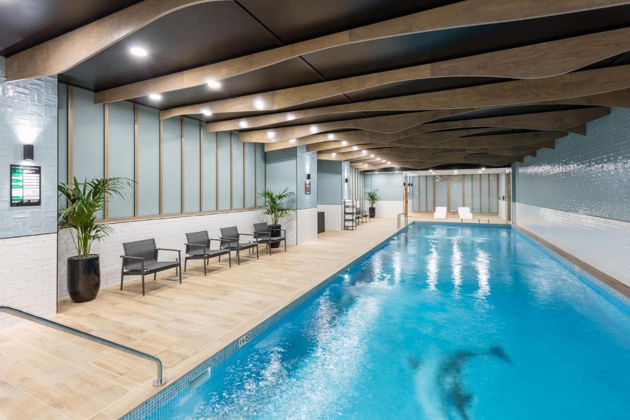 Meriton Suites Sussex Street, Sydney Eksteriør billede The photo shows an indoor swimming pool area. The pool has clear blue water and is flanked by a wooden deck. There are several lounge chairs arranged along one side of the pool. The ceiling features a modern design with wooden beams and recessed ligh