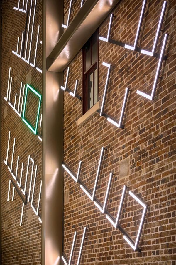 Meriton Suites Sussex Street, Sydney Eksteriør billede The photo shows a brick wall featuring a design made up of linear light fixtures. These light strips create geometric shapes and patterns, with some sections illuminated in white and one section highlighted in green. The combination of the brick text