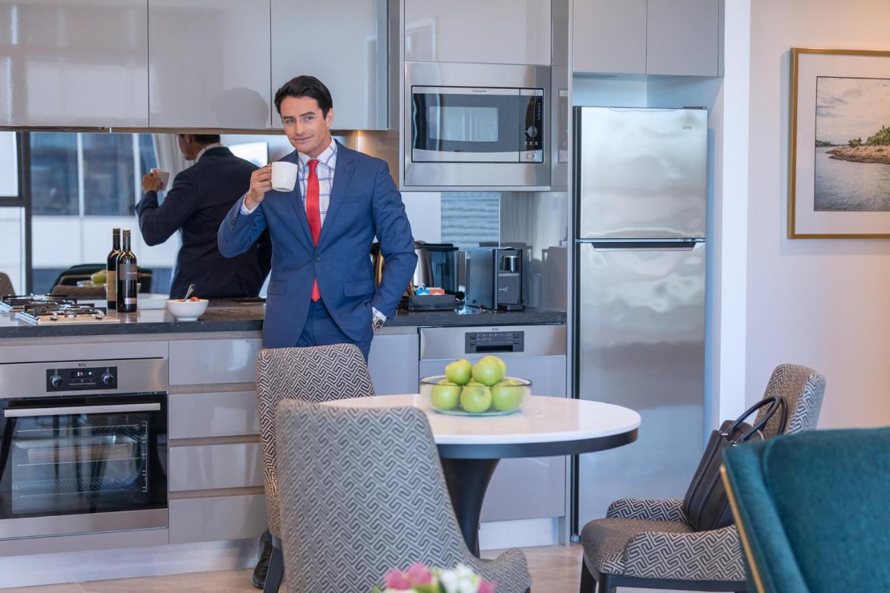 Meriton Suites Sussex Street, Sydney Eksteriør billede The photo depicts a well-dressed man in a suit standing in a modern kitchen. He is holding a cup, possibly of coffee, and appears to be looking towards the camera. The kitchen features contemporary appliances, including a microwave and refrigerator. 