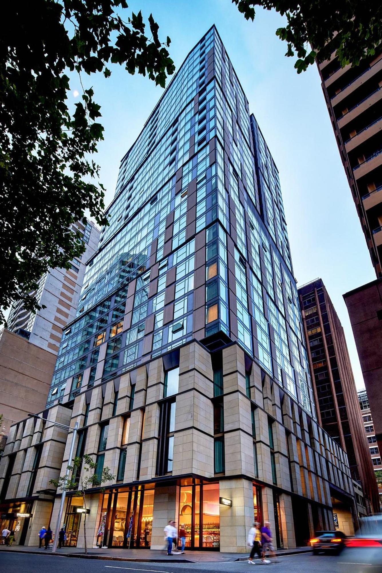 Meriton Suites Sussex Street, Sydney Eksteriør billede The photo shows a modern high-rise building with a sleek, glass facade reflecting its surroundings. The structure features a combination of glass and concrete materials, with multiple stories visible. The lower part of the building has a sturdy, text