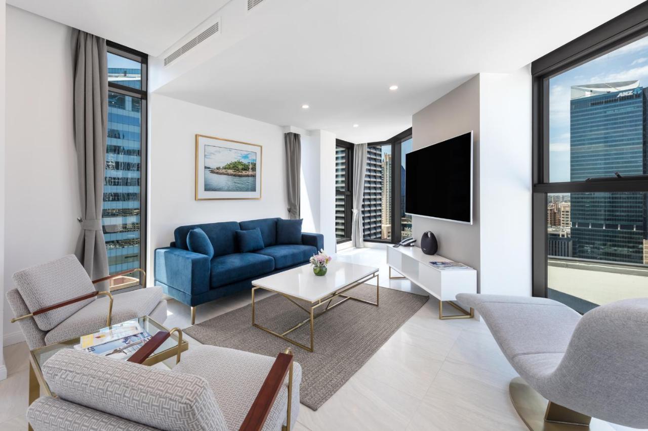 Meriton Suites Sussex Street, Sydney Eksteriør billede The photo shows a modern living room featuring large windows that allow plenty of natural light. The room is elegantly designed with a blue sofa, a light-colored coffee table, and two armchairs. There’s a flat-screen TV mounted on the wall, and a pie